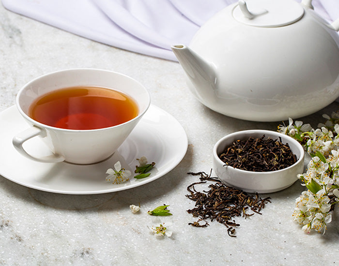 A white teacup with tea, beside a teapot, dried tea leaves, and flowers.