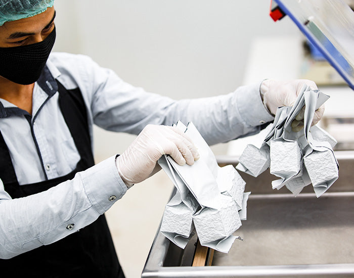 Worker with gloves and face mask handling sealed white packages in a manufacturing setting.