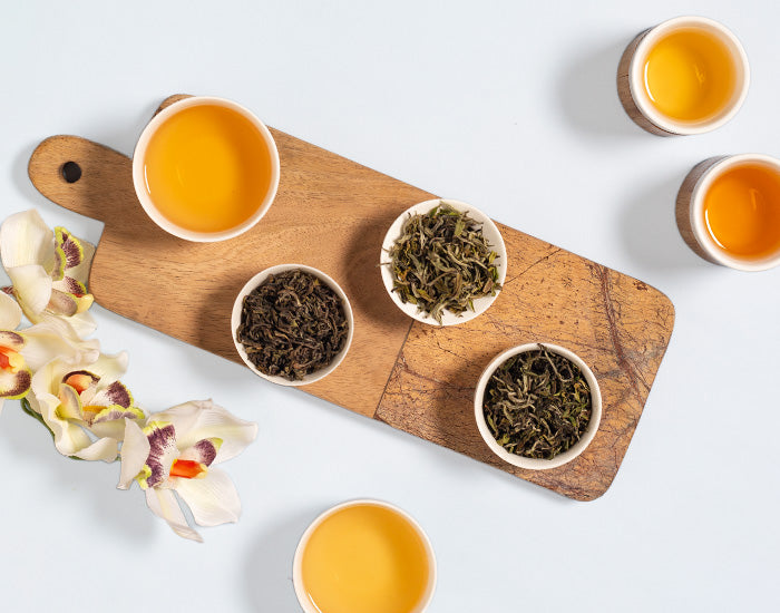 Tea cups and bowls with loose leaf tea on a wooden board, accompanied by flowers.